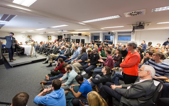 A large room full of about 100 people, watching a man in a suit giving a presentation and pointing to the screen. In the background cameras.
