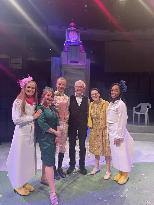 Five actors dressed in costumes for the play Ladies' Day in front of a clock tour with Tony Christie in the middle