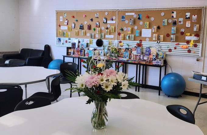 A classroom with tables and chairs. An assortment of books that include novels, creative nonfiction, magazines, and children's picture books, are displayed on tables against a wall. 