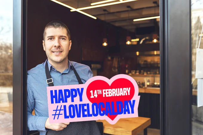 Image depicts a shopkeeper holding a sign which reads ‘Happy #LoveLocalDay 14TH February’