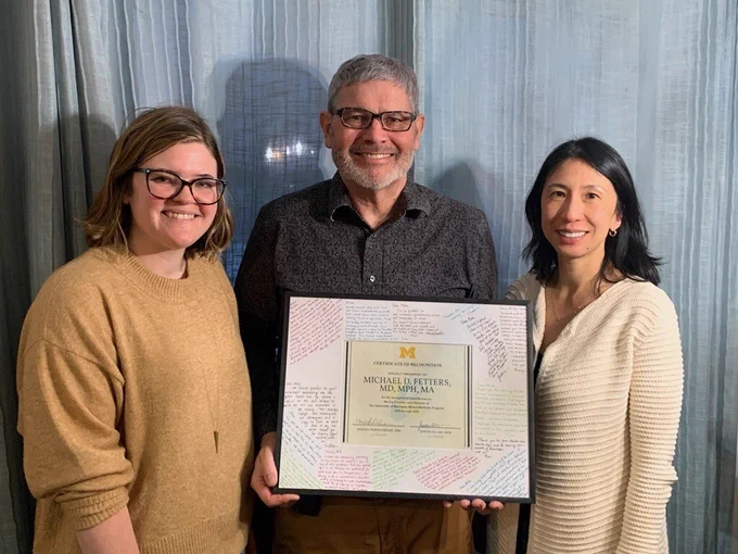 photo of three people with the person in the middle holding a framed certificate. 