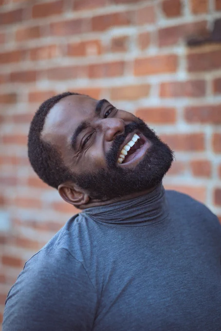 Jerron, a delicious chocolate brother, laughs against a brick wall wearing a grey turtleneck. Sure, it’s a headshot but isn’t it also joy?
