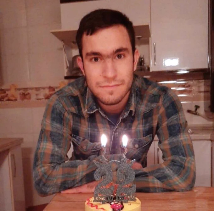Mohammad sitting at a kitchen table with his 22nd birthday cake in front of him. He has a long face and short dark brown hair, a long forehead, brown eyebrows and eyes, an average-size straight nose, thin lips, and a very short, nicely trimmed, full beard. He's wearing a plaid (gray and dark orange) shirt and his forearms are gently crossed and resting on the table. The table is light brown and wooden, and the background includes kitchen cupboards and walls. 