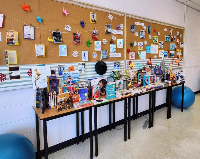 An assortment of books that include novels, creative nonfiction, magazines, and children's picture books, displayed on multiple tables in a classroom. A bulletin board behind it shows covers of more books.