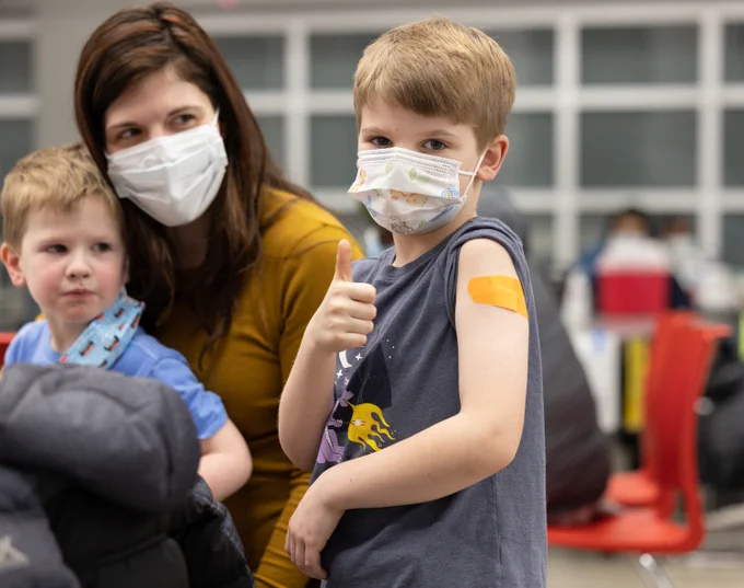 Mom with a toddler stands by a masked boy who has a bandage on his arm and giving thumbs up.