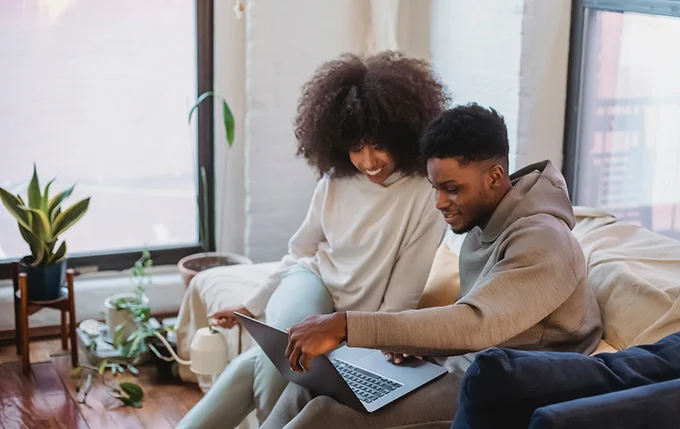 A couple sits on a couch in a bright room with plants by the window. They are smiling and looking at a laptop.