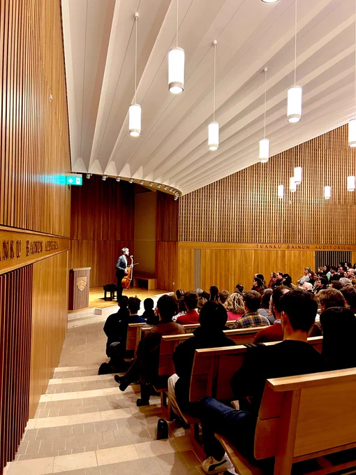 Cellist Vladimir Waltham stands on stage with his cello in the Sultan Nazrin Shah Centre, Worcester College Oxford, addressing the large audience.