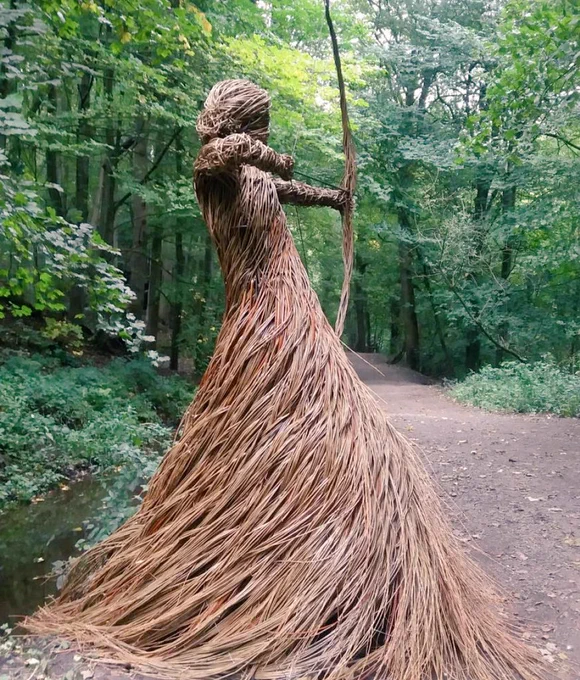 Photograph of a scuplture in a woodland setting, the sculpture features a figure in a long dress in a pose as if about to release an arrow from a bow, facing away from the viewer, the figure looks female and is constructed with many brown willow strands twisted into shape, it stands on a path surrounded by trees