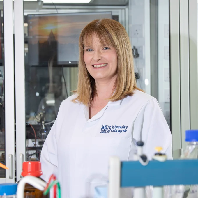 Dr Nicola Bell. She is standing in a science lab and wearing a white lab coat that has the University of Glasgow logo on it. 