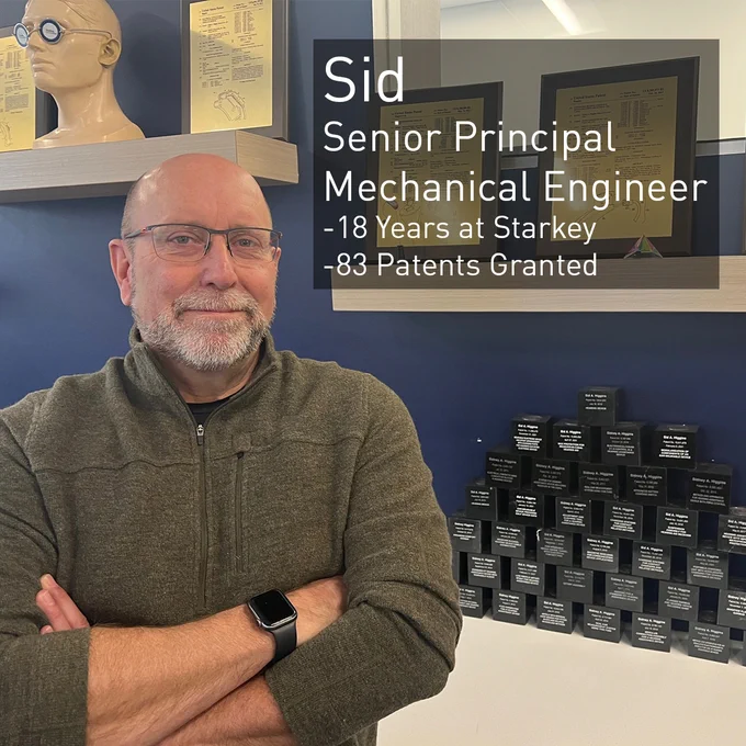 Sid, a Senior Principal Mechanical Engineer, stands infront of a large display of awards and smiles. He's been with Starkey for 18 years and has 83 patents granted.