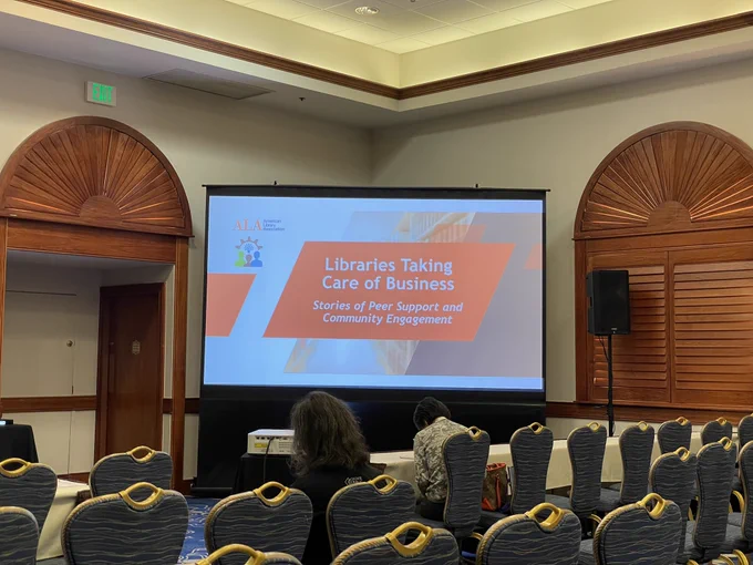 Powerpoint in the front of a ballroom, with the title “Libraries Taking Care of Business: Stories of Peer Support and Community Engagement”