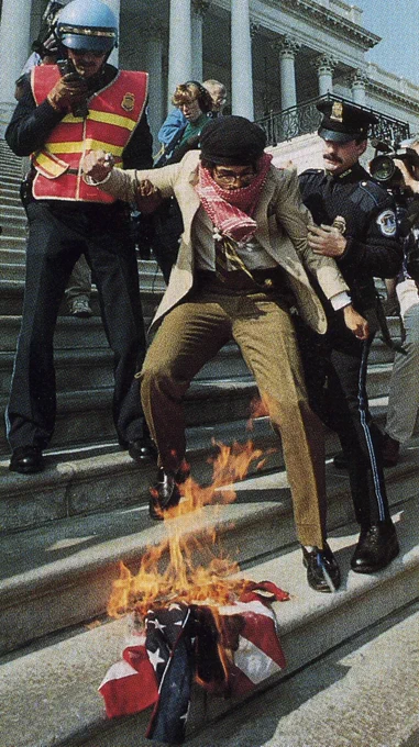 Dread Scott burning a U.S. flag on the steps of the US Capitol, 1989. He is held on each side by police officers. 