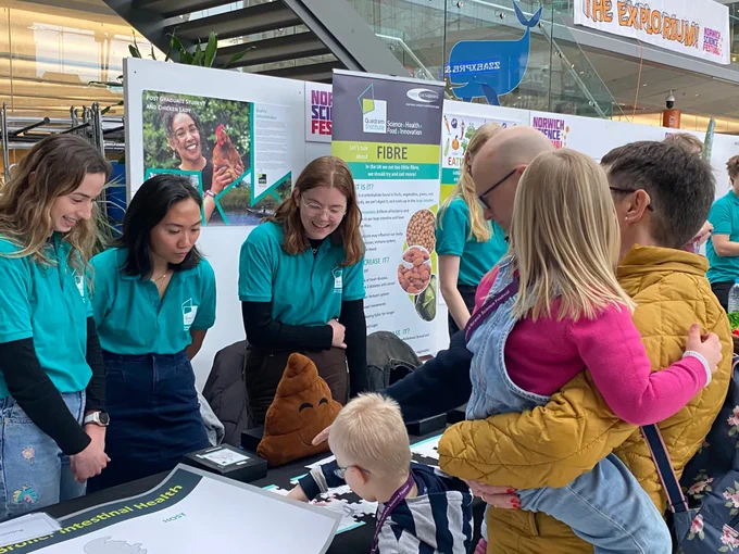 Three young women in turquoise shirts at a stall at the Norwich science festival including a poo emoji cushion. Members of the public looking interested. 