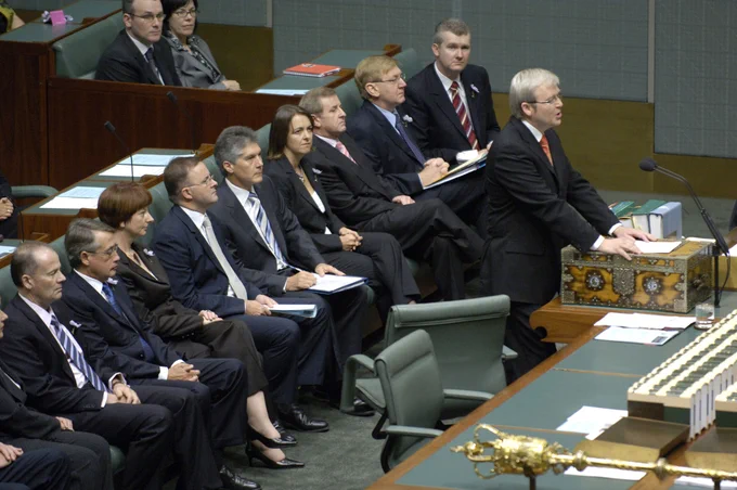 Former Prime Minister Kevin Rudd stands at the dispatch box in the House of Representatives Chamber to deliver the National Apology to Australia’s Indigenous Peoples. He wears a black suit, red tie and glasses. Behind him are other members of parliament seated on green benches, including Deputy Prime Minister Julia Gillard and Foreign Minister Stephen Smith. They are dressed in business attire.