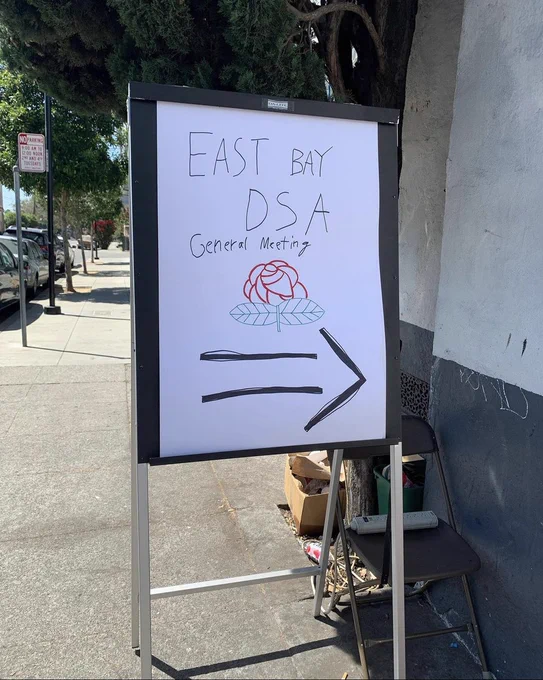 A whiteboard on the sidewalk with text reading "East Bay DSA General Meeting." Below is a drawing of a rose. At the bottom is a large arrow pointing to the right.