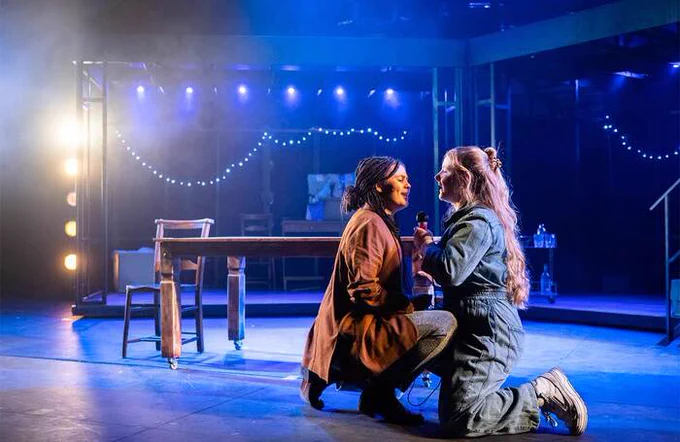 Two performers on stage crouched down looking at each other singing to each other 