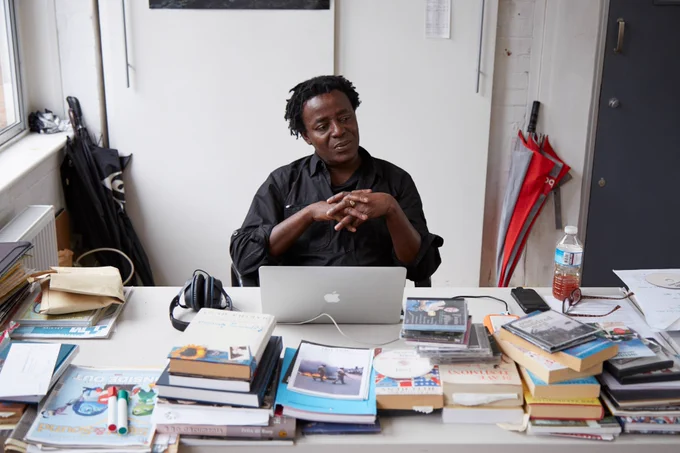 A man wearing a black shirt sits at a desk with many books, pens and a laptop.
