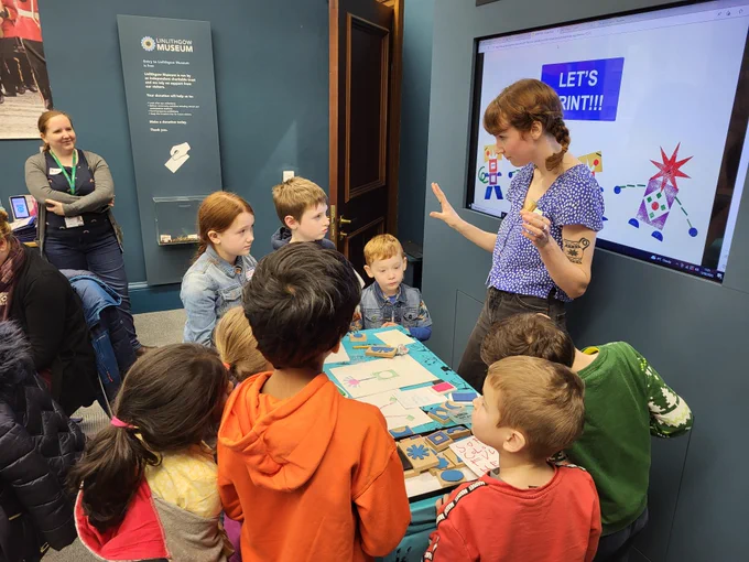 A group of children is gathered round an artist who is explaining what the workshop activity will be. There is a projector in the background reading 'let's print' and everyone is silently listening
