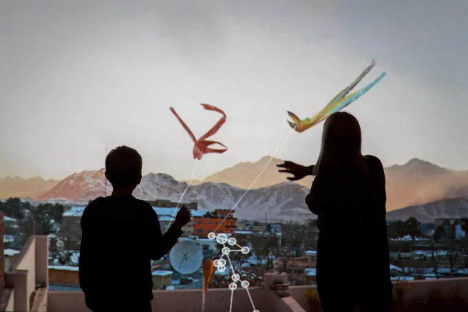 Two people looking at a projection on a wall of a mountain area and two kites flying in the wind.
