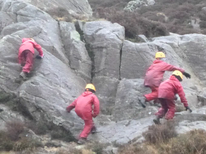 Pupils in helmets and waterproofs scrambling on a rock face in the mountains