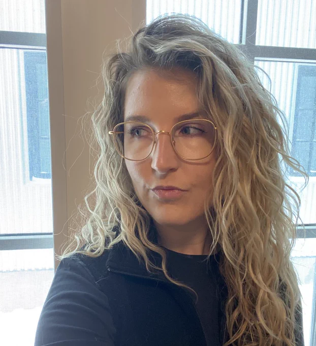 Image description courtesy of author: Photograph of Amy, a white woman with green eyes and long, wavy blonde hair. In this picture Amy is standing in front of a multi-paned window. She is wearing gold wire frame glasses, a black long-sleeved t-shirt, and a black fleece vest. She is smiling softly and looking towards the left of the image.