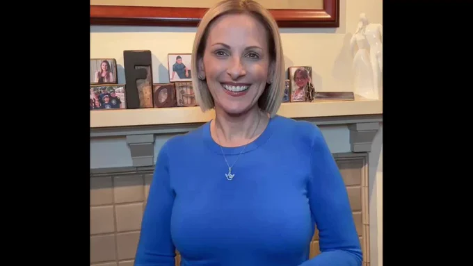 Actress Marlee Matlin is sporting short blonde hair, wearing a blue blouse and a pendant representing the words "I Love You" in ASL around her neck. She is standing in front of a fireplace with pictures of her children on the mantle. 