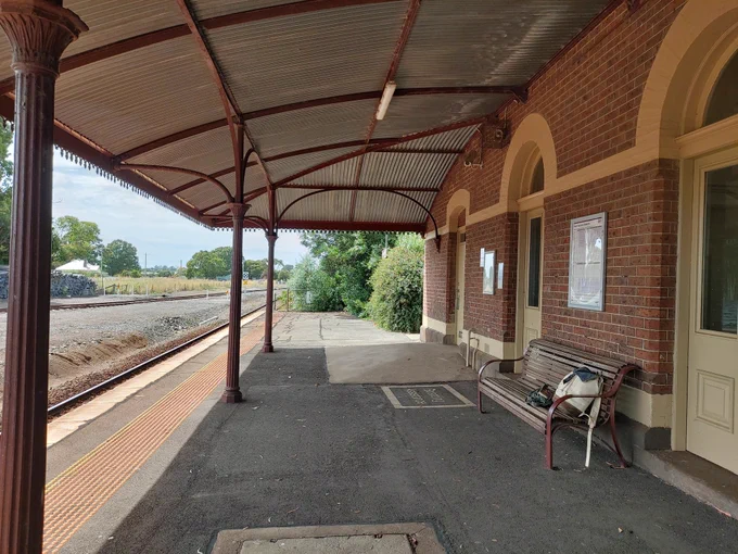An old country train station platform, tracks disappearing into the distance. A canvas rucksack sits on a bench, unattended. 