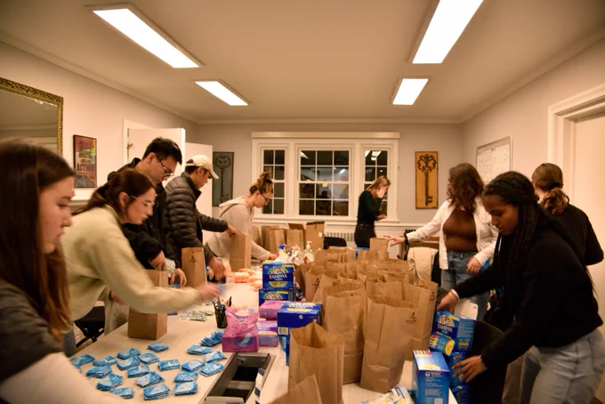 People stand around a table. On the table there are paper bags, menstrual pads, and boxes of tampons "Tampax". The people are picking up boxes and pads and placing menstrual products into the brown paper bags. 