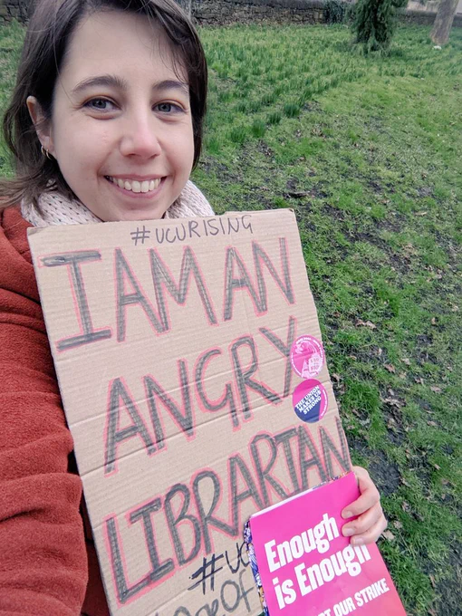 A university staff member on strike, holding a sign saying I am an angry librarian, with daffodil shoots in the background 