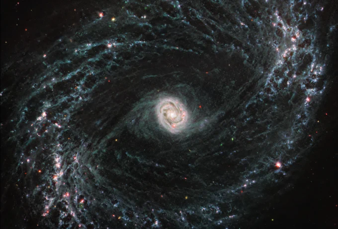 A large galaxy takes up the entirety of the image. The core is mostly bright white, but there are also swirling, detailed structures that resemble water circling a drain. There is white and light blue colored dust that emanates from the core’s center, but it is tightly contained to the core. The rings are wispy and highlight filaments of dust around cavernous black bubbles. The dust in the outer rings contains dots that are navy blue, pinkish, reddish, and white.