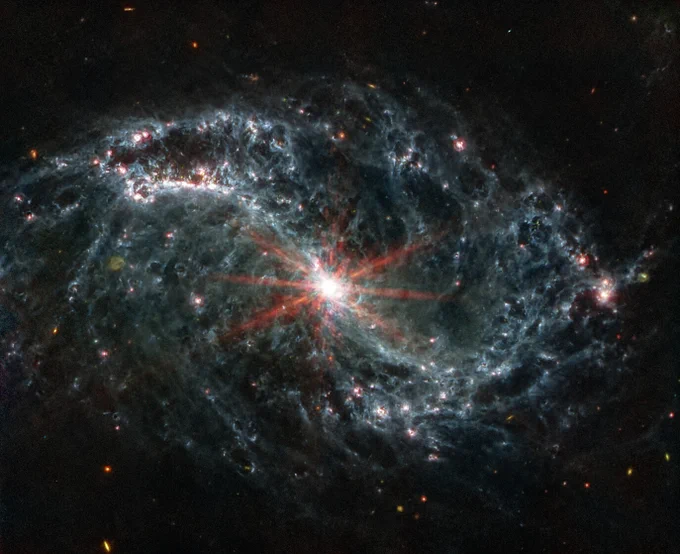 A large galaxy takes up the entirety of the image. Six reddish diffraction spikes extend from the core. The spiral arms of the galaxy outstretch to the upper left and the lower right. The arms are wispy and highlight filaments of dust around cavernous black bubbles. That dust in the outer rings contains diffuse dots that are navy blue, pinkish, reddish, and white.