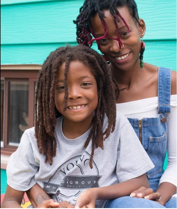 A Black woman (Tina Opalke) sits smiling at the camera. Her hair is in dreadlocks and she is wearing denim coveralls and a white shirt. Her young daughter is sitting in her lap and is also smiling. Her hair is in dreadlocks and she is wearing a t-shirt. A blue clapboard house is in the background. 