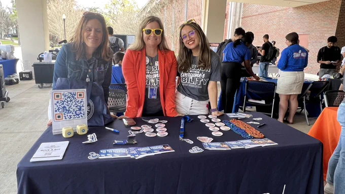 Three disability resource center staff wearing “inclusion is within everyone’s ability” t-shirts stand behind their table at a giving day event.
