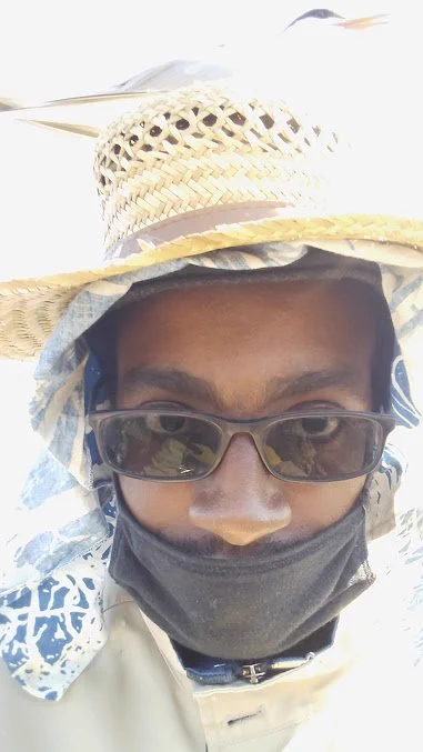 A selfie of Adé, a young black man wearing a mask and two hats, featuring a common tern sitting on his head.