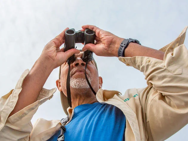 Photograph of Derrick Z. Jackson – a older Black man with a white goatee – taken from below as he looks through binoculars, overcast blue sky behind him.
