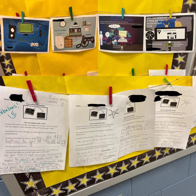 Eight pieces of paper hung on a bulletin board in a classroom. The top four show drawings made by students to illustrate the concept of Input, Process, Storage, and Output. The bottom four papers show students’ written descriptions of how their microbits sent radio signals to each other.