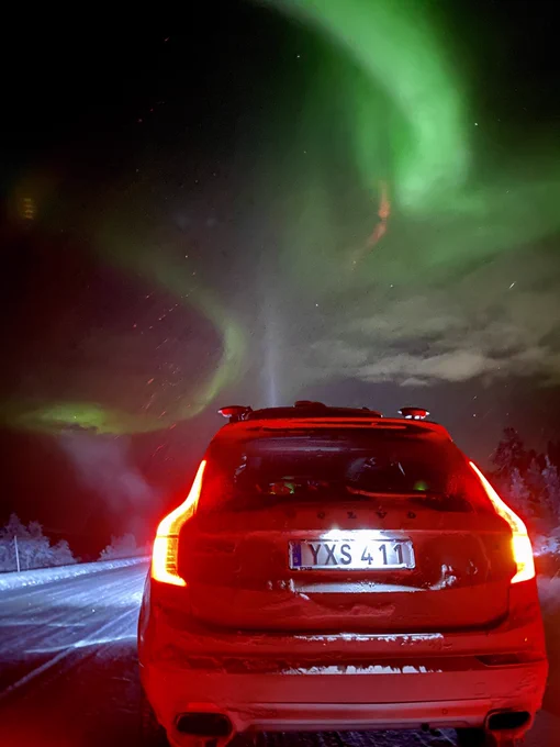 White XC90 Volvo on the side of a snowy road, under Northern Lights.
