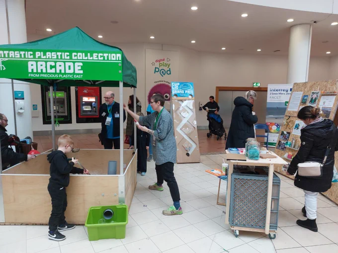 Two people have little pickers in their hands, and are collecting plastic cups from a large wooden container inclosed in a gazebo. The top of the gazebo is green and has the words ‘fantastic, plastic collection, arcade, from Precious Plastic East’ in white. Beyond the gazebo, people are looking at the exhibition’s display boards. These have images on, including a green and pink illustration of David Attenborough.