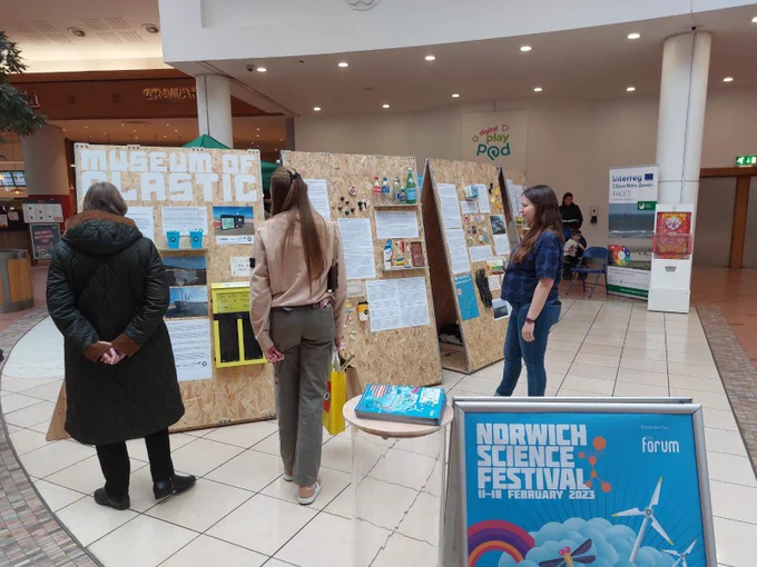 In the foreground is the top of an a-frame board showing a Norwich Science Festival poster. The poster is blue, with the festival’s logo in white. Beyond this, in the background are three people looking at the exhibition’s boards. Each of the display boards have posters and images attached. The middle board has plastic bottles lined up on a plinth.