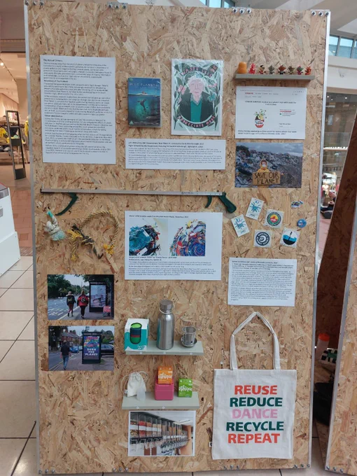 A display board shows images and items - all with a theme of reducing plastic consumption. One image is of a child in front of a large heap of rubbish, they are holding a placard that says ‘Save our planet’. There is a white tote bag with the words ‘reuse, reduce, dance, recycle, repeat’ in green, pink and orange. There is also a little picker alongside some rope, metal reusable coffee cups, a Blue Planet DVD case and an illustration of David Attenborough.