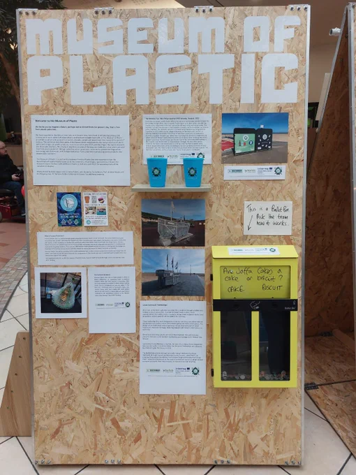 A display board with the words ‘Museum of Plastic’ at the top in large white letters. Underneath this are two plastic cups on a plinth and photos of installations and pieces of art made of reused plastic items; such as a Christmas tree entirely made of plastic bottles.