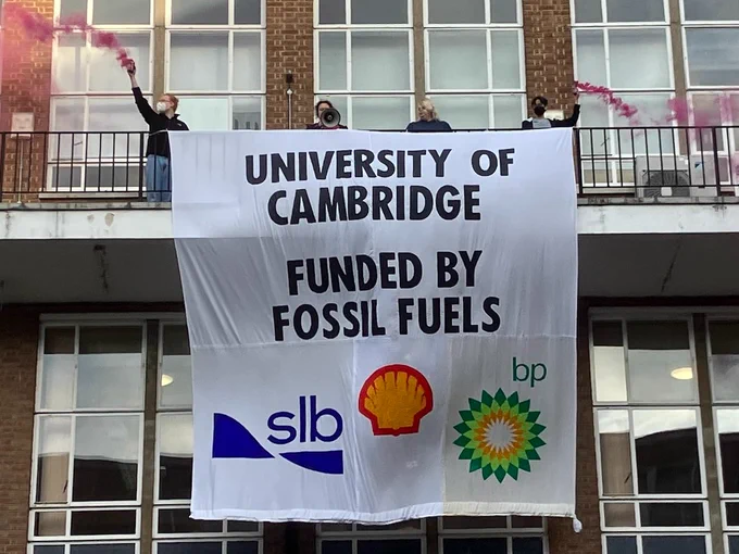 Four rebels on a balcony holding a banner which reads "University of Cambridge funded by fossil fuels" and displays the logos of SLB (Schlumberger), Shell, and BP.