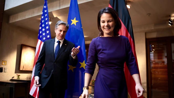 Secretary Blinken gestures with his hand following a photo with German Foreign Minister Baerbock. U.S. , European and German flags sit behind them.