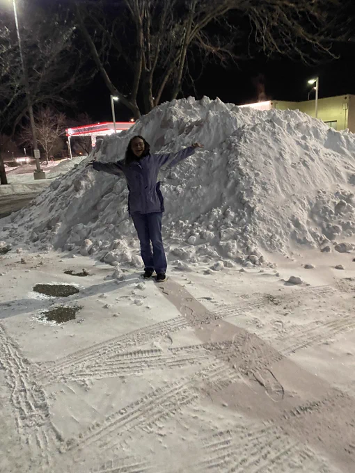 Smiling woman (me) standing in front of a snow pile larger than her with her arms spread out. She’s thinking about how you don’t find anything like this in Florida.