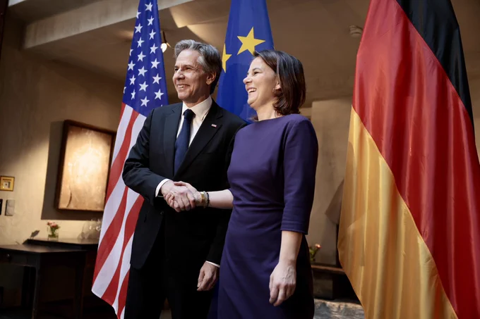 Secretary Blinken and Foreign Minister Baerbock shake hands and pose for a photo together while standing in front of U.S., European and German flags.