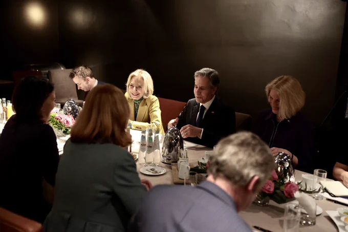 Secretary Blinken, Foreign Minister Baerbock and advisors sit across from one another at a conference table flanked with flowers, glasses and china.