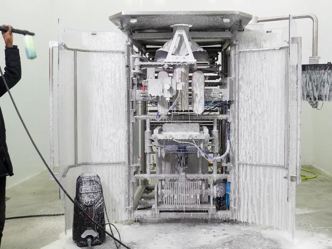 A photograph of WeighPack's VF-1200 continuous motion vertical bagger. Its frame has been opened to reveal the inner machinery, and an operator, standing off-camera, is spraying the interior with soapy water. 