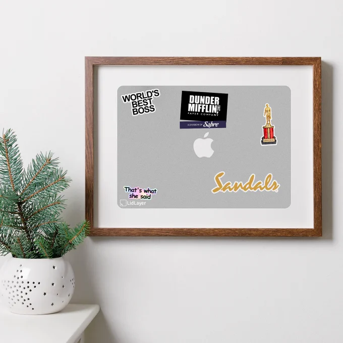 A framed LidLayer of a silver MacBook Pro, with stickers associated with Michael Scott, such as 'Worlds Best Boss', the Dunder Mifflin logo, and "that's what she said"