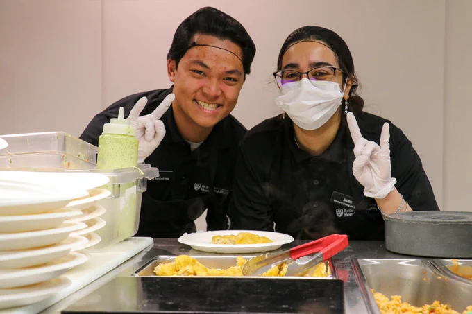 Two student workers wearing black uniforms, hair nets and gloves smile at the camera and hold up the hook ‘em hand sign. Photo courtesy of Ramces Luna.
