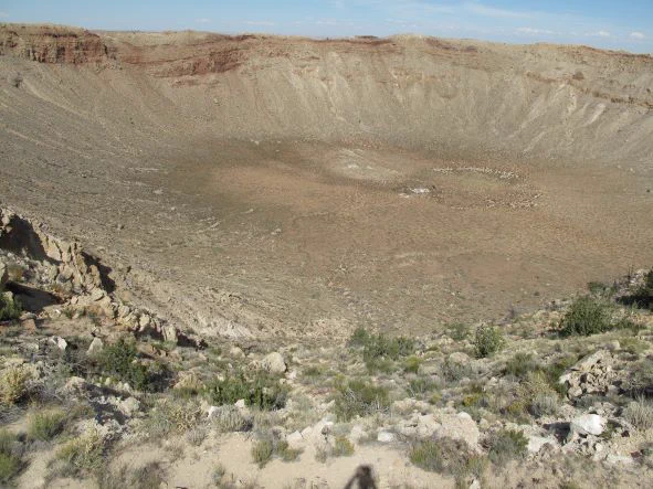 Large circular meteor crater in Arizona.  The ground in the crater is brown and rocky.  Green brush surrounds the crater.